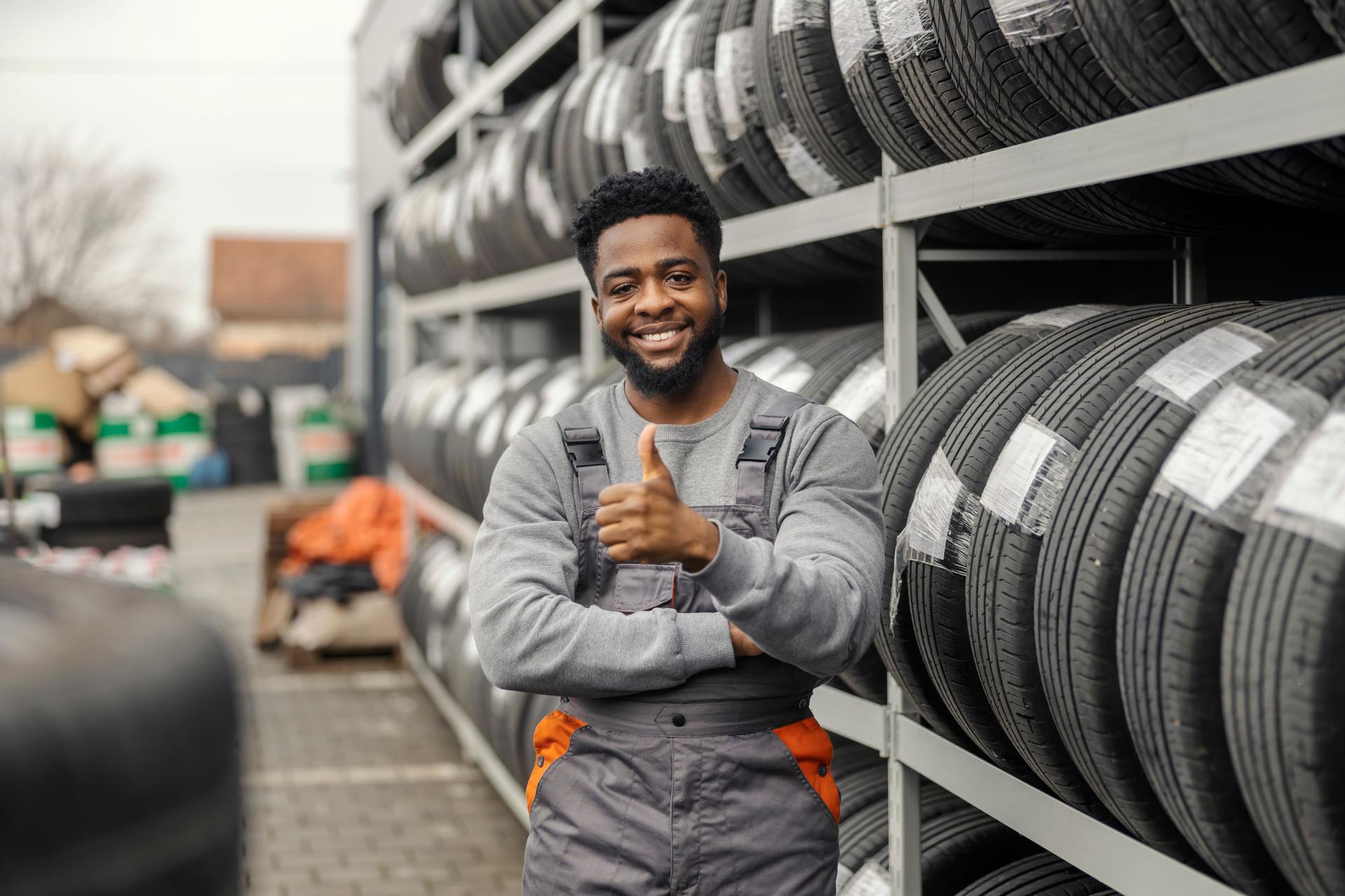 Portrait of african american technician standing near piles of tire and giving thumbs up at camera.